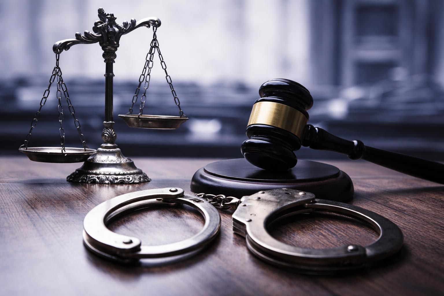 The image shows a judicial gavel, a set of handcuffs, and a scale of justice, all resting on a wooden surface with a backdrop of a courtroom setting.