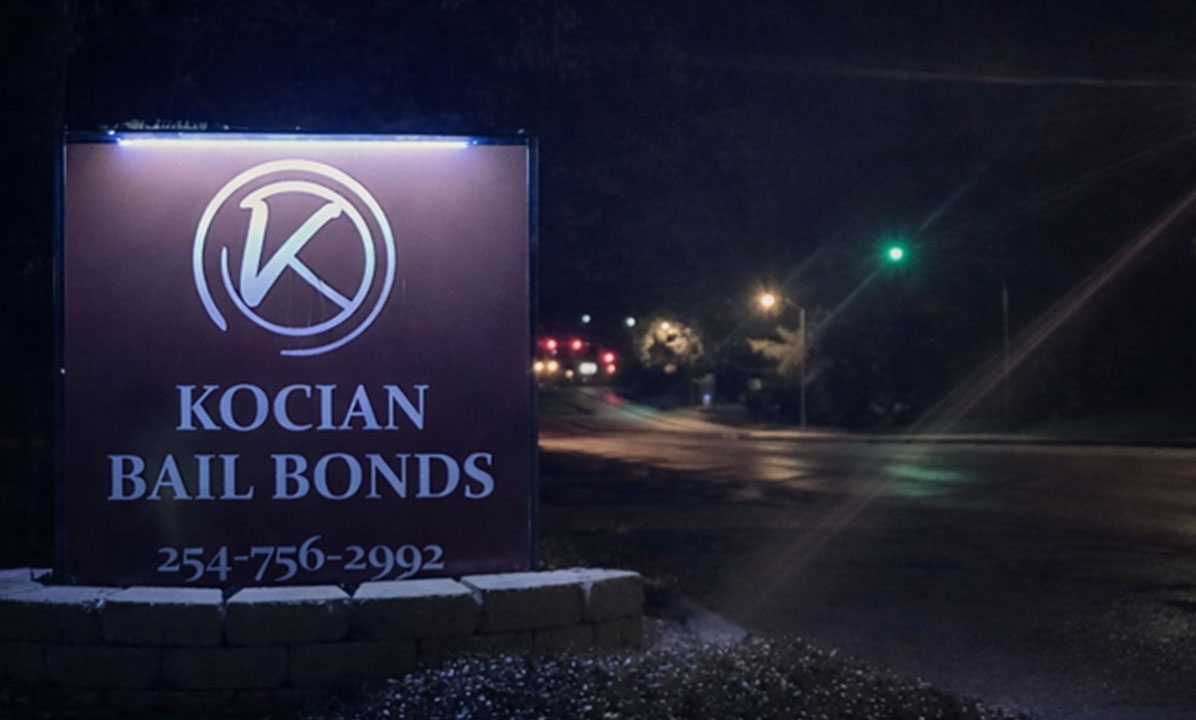 The image shows a nighttime view of a sign for Kocian Bail Bonds with a lit-up logo, located on a street corner at night.